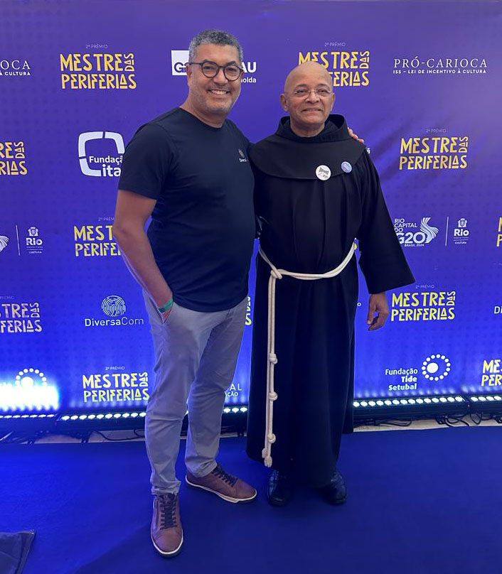 Marcelo Moreira, Sócio-Diretor da DiversaCom e Frei David em pose para foto com um banner do evento Mestre das Periferias atrás deles