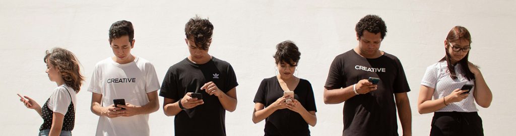 Seis jovens em pé, lado a lado, utilizando seus smartphones, diante de uma parede clara. Dois deles vestem camisetas com a palavra “CREATIVE”.